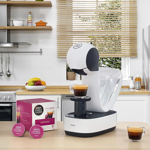Cafetera Dolce Gusto Infinissima preparando espresso en cocina moderna Cafetera Dolce Gusto Infinissima blanca preparando un espresso en una cocina moderna, con cápsulas y caja de café a su lado.