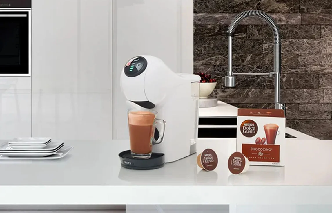 Cafetera Dolce Gusto Genio S blanca preparando Chococino en cocina moderna Cafetera Genio S Dolce Gusto blanca en cocina moderna, sirviendo un Chococino. Acompañada de cápsulas y empaque, este modelo Genio S Plus destaca por su diseño elegante y funcional.